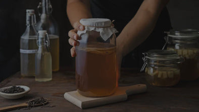 A glass jar of homemade kombucha tea with a scoby, covered with a cloth lid, placed on a wooden board, surrounded by tea leaves and ingredients, representing a natural, organic, probiotic-rich fermented drink for gut health and wellness.