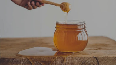 A honey dipper lifts golden honey out of a glass jar, with a single stream of honey dripping back into the jar and a small puddle of honey on the rustic wooden table.
