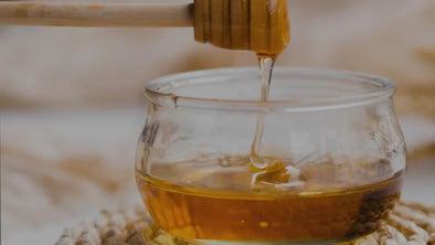  A wooden honey dipper is held over a small glass bowl, with a thin stream of golden honey dripping into the bowl. The background is a soft, light-colored blur.
