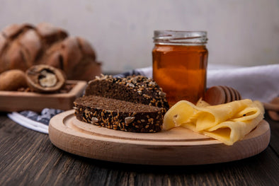 Wooden board with sliced dark seed bread, melted cheese, a jar of honey, and walnuts on a dark wooden surface with bread rolls in the background.