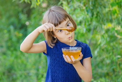 A young girl in a blue dress holds a glass bowl of honey while playfully lifting honey with a wooden dipper. She looks at the dripping honey with curiosity. The background is lush and green, suggesting an outdoor setting.