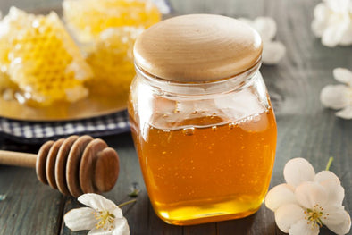 A glass jar filled with golden honey, topped with a wooden lid, placed on a rustic wooden surface. Nearby are pieces of honeycomb on a plate, a wooden honey dipper, and scattered white flowers, with a checkered cloth in the background.