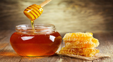 A jar of honey sits on a wooden surface next to a piece of honeycomb, with a honey dipper drizzling honey into the jar.