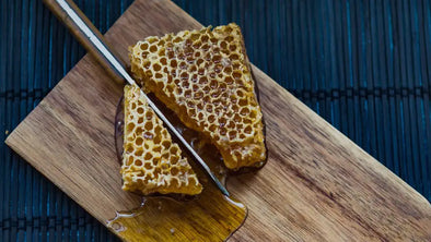 A close-up shot of two thick, golden chunks of raw honey comb resting on a wooden cutting board. A small knife is slicing into one of the pieces, and pure, viscous honey is dripping and pooling onto the board below.