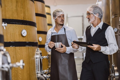 Two workers honey production facility wearing hairnets and discussing quality control. One holds a tablet while the other holds a measuring cup, standing near large wooden fermentation tanks with gauges.