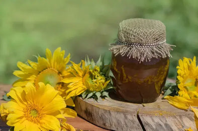 Jar of honey with a burlap cover placed on a wooden surface, surrounded by bright yellow sunflowers in an outdoor setting.