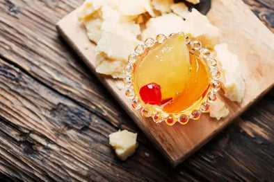 A close-up of a small glass dish containing a pear slice in a golden syrup with a red cherry, placed on a wooden board surrounded by chunks of Parmesan cheese.