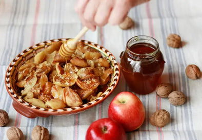 A hand drizzles honey from a wooden dipper over a decorative clay bowl filled with baked apple slices, placed on a striped tablecloth. Nearby are a jar of honey, two red apples, and scattered walnuts.