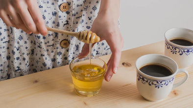 A person uses a wooden honey dipper to drizzle golden honey into a small glass bowl on a light wooden table, next to two mugs of black coffee.