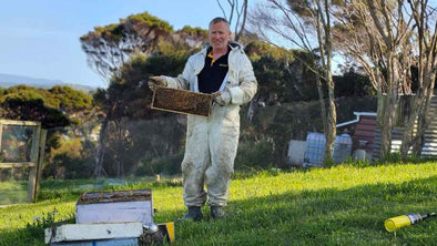 A beekeeper in a white protective suit holds a honeycomb frame with bees, standing on a grassy field with beehives and trees in the background, under a clear blue sky.