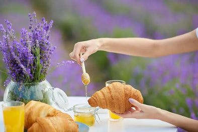 A person drizzling honey from a wooden dipper onto a croissant, with more croissants, a jar of honey, a glass of orange juice, and a vase of lavender flowers on a table, set against a backdrop of a lavender field.