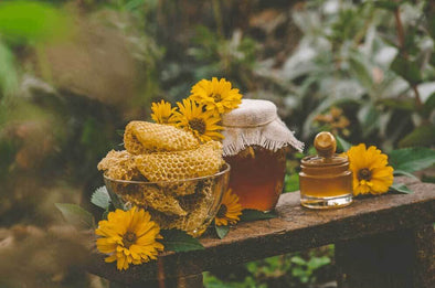 A jar of honey sits on a wooden board next to honeycomb, a honey dipper, and lavender.