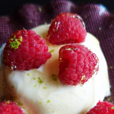 A close-up of a creamy white dessert topped with fresh raspberries and small pieces of lime zest, served on a dark purple napkin.