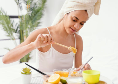A woman with a towel wrapped around her head applies honey to a homemade skincare mixture. She is seated at a table with natural ingredients like lemon and yogurt, creating a DIY beauty treatment. The setting is bright and relaxing.