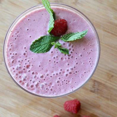 A vibrant pink smoothie in a glass, garnished with fresh mint leaves and a raspberry on top, placed on a wooden surface with additional raspberries scattered around.