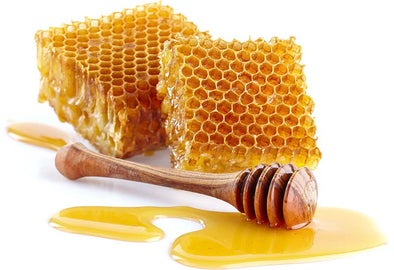Two pieces of honeycomb with honey dripping onto a white surface, accompanied by a wooden honey dipper with honey on it.