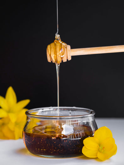 Vendor's stall displaying a jar of raw honey with a dipper, fresh produce, and colorful flowers under a clear sky.
