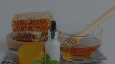 A glass jar of golden amber honey with a wooden dipper, surrounded by a rustic wooden spoon and bowl containing beeswax comb and natural skincare balm, featuring mint leaves, representing a pure organic remedy for wellness and beauty.