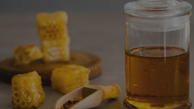 A glass jar filled with golden honey next to a wooden scoop with bee pollen, accompanied by pieces of honeycomb on a wooden base, set against a blurred background.