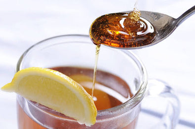 A glass cup of hot tea with a slice of lemon on the rim, while a spoon drizzles golden honey into the tea.