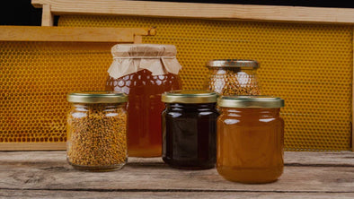 Vendor's stall displaying a jar of raw honey with a dipper, fresh produce, and colorful flowers under a clear sky.