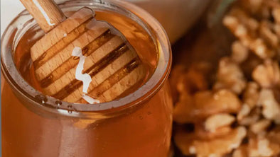 A close-up shot of a wooden honey dipper submerged in a glass jar filled with rich, golden liquid honey. To the right, a pile of shelled walnuts is blurred in the background.