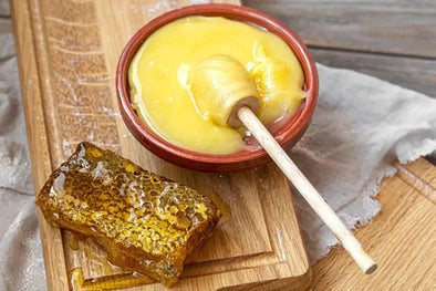 A wooden board with a piece of honeycomb and a red clay pot filled with honey, a wooden dipper resting in the pot, and a light cloth in the background.