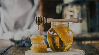 A glass jar filled with honey and a large piece of honeycomb sits on a wooden surface. A wooden honey dipper rests across the top of the jar.