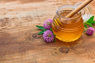 A glass jar of golden honey with a wooden dipper, surrounded by purple clover flowers and green leaves on a rustic wooden surface, with the jar's lid nearby.