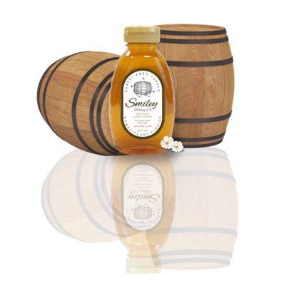 Two glass jars of golden honey, each containing a large piece of intact, yellow-orange honeycomb, are displayed on a wooden shelf.