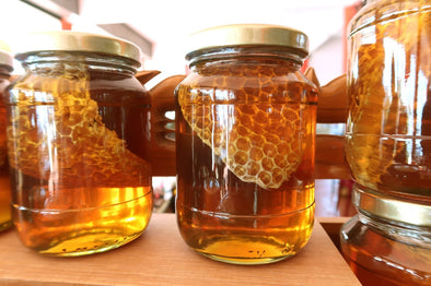 Two glass jars of golden honey, each containing a large piece of intact, yellow-orange honeycomb, are displayed on a wooden shelf.